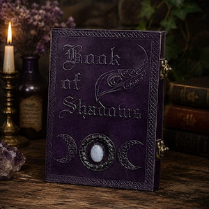 Op de afbeelding: Een rechthoekig paars boek met de tekst Book of Shadows in reliëf op de voorkant. De kaft heeft een centrale ovale maansteen cabochon omringd door een donkere metalen ring, geflankeerd door twee symbolen van een wassende maan.