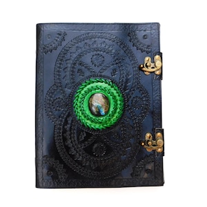 May include: A black leather journal featuring intricate embossed circular patterns across the cover. A bright green braided border surrounds a central iridescent oval gemstone. Two brass-colored metal latches secure the side of the book.