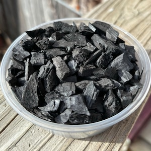 May include: A clear plastic container filled with black charcoal pieces. The charcoal is broken into small, irregular pieces.