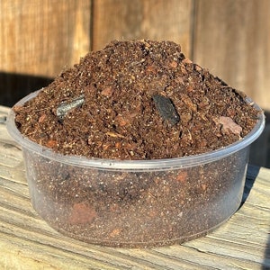 May include: A close-up of a brown potting mix in a clear plastic container. The mix contains small pieces of bark and other organic matter.