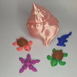 May include: A large, pearlescent pink conch shell container accompanied by four small, articulated plastic figures: a pink and brown turtle, a green and brown turtle, a blue hammerhead shark, and a purple and blue spotted starfish.