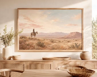 Puede incluir: Una impresión de arte mural de paisaje enmarcada que representa a una persona a caballo en un entorno desértico. La escena muestra hierbas secas, cactus y mesetas distantes bajo un cielo suave de color rosa pálido y azul. Marco de madera clara.