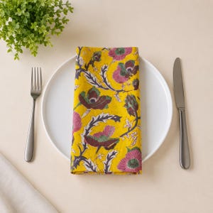 Puede incluir: Una servilleta de tela amarilla doblada con un patrón floral repetitivo en tonos rosa, verde oscuro y negro. La servilleta está colocada sobre un plato de cerámica blanca, acompañada por un tenedor de metal plateado a la izquierda y un cuchillo a la derecha.