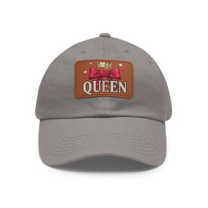 May include: A grey baseball cap featuring a rectangular brown patch on the front. The patch displays a gold crown above a red bow with a heart center, and the word QUEEN in white capital letters with a dark outline, surrounded by small hearts.