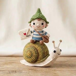 Op de afbeelding: Een gehaakt amigurumi-figuurtje van een klein personage met een groene puntmuts en een blauw-wit gestreept shirt, rijdend op een grote slak met een groen en bruin spiraalvormig huisje. Het personage houdt een witte envelop vast met een rood hartje en het woord MAIL.