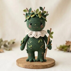 May include: A dark green crocheted doll with a round head, large black eyes, and rosy cheeks. It wears a white scalloped collar and has small green leaves and brown twigs attached to its head and body. It holds a thin brown wooden staff.