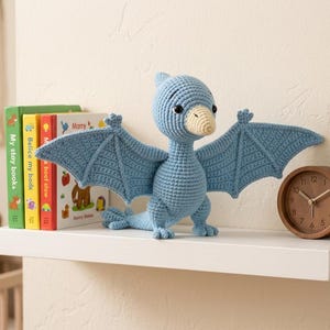 May include: A light blue crocheted pterodactyl toy with a cream-colored beak and black safety eyes. The stuffed creature has outstretched wings, a long tail, and stands on two feet. It is positioned between three colorful books and a round wooden clock.
