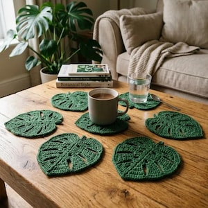 May include: A set of six dark green crochet coasters shaped like monstera leaves. Each coaster features a detailed leaf vein pattern with cutouts. One coaster supports a light grey ceramic mug filled with a brown beverage, while another holds a glass of water.