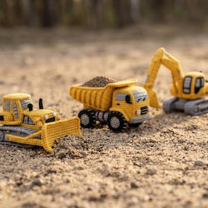 Könnte beinhalten: Drei gehäkelte Baufahrzeuge aus gelbem und grauem Garn. Ein Bulldozer mit Frontschild, ein mit kleinen grauen Steinen beladener Muldenkipper und ein Bagger mit beweglichem Arm sind auf einer sandigen, kiesartigen Oberfläche angeordnet.