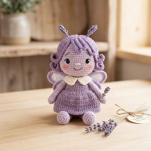 Può includere: Una bambola a forma di fata all'uncinetto fatta a mano, nei toni del viola, con colletto e ali bianche. La bambola ha un viso sorridente con guance rosee e tiene in mano un rametto di lavanda. Un piccolo mazzetto di lavanda e un'etichetta sono accanto alla bambola.