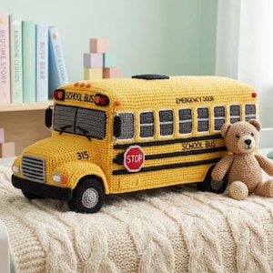 May include: A yellow crocheted school bus toy with black accents, featuring the text SCHOOL BUS and STOP on the side, and EMERGENCY DOOR on the back. A small brown crocheted teddy bear sits next to the bus on a cream-colored knitted blanket.