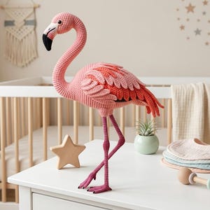 May include: A crochet flamingo toy featuring a long, curved neck, a beak with black and white colouring, and a body with layered pink and orange yarn feathers. The flamingo stands on two long, thin, dark pink legs with detailed feet.