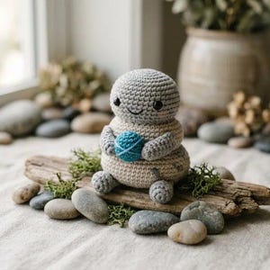 May include: A small, round, crocheted creature with a light gray head and a beige body. It has black bead eyes, a stitched smile, and pink cheek accents. The figure holds a small, textured, bright blue ball of yarn between its two arms.