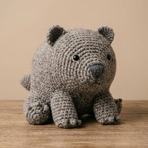 May include: A grey crocheted stuffed animal shaped like a wombat with a round body, short limbs, small rounded ears, and a dark grey stitched nose. The toy features a textured yarn surface and two small black plastic eyes set into the head.