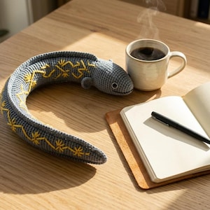 May include: A grey crocheted eel-shaped plush toy featuring yellow embroidered star-like patterns along its curved body, positioned next to a ceramic mug filled with a dark beverage and an open notebook with a black pen resting on its pages.