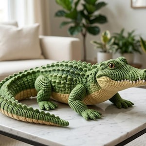 May include: A handcrafted crochet alligator toy featuring a textured green back with raised scales and a smooth light beige underbelly. The plush figure has a long curved tail, four short legs with defined toes, and a wide snout with visible white teeth.