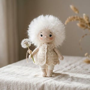 以下が含まれることがあります： ふわふわの白い髪とクリーム色の衣装を着た、手作りのかぎ針編みの人形。人形はタンポポを持っています。人形はベージュのリネンのテーブルクロスの上に立っています。背景には、ドライフラワーが入った花瓶があります。