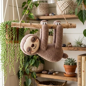Peut inclure: Un paresseux en crochet marron avec un visage crème, des yeux fermés et un petit sourire, suspendu à une branche en bois clair. Le paresseux a de longs bras avec des griffes blanches et est accroché par des cordes fines parmi des plantes.