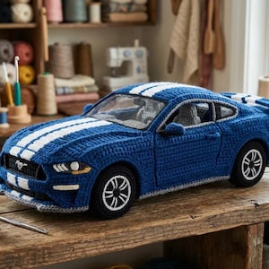 Pode incluir: Um modelo de carro em crochê azul com duas faixas de corrida brancas no capô e no teto. O veículo possui rodas pretas com aros brancos, uma grade frontal com um pequeno emblema e assentos internos visíveis feitos de fio.