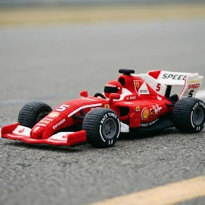 以下が含まれることがあります： 赤と白のクロシェ編みのF1レースカーのおもちゃ。ノーズとリアウィングに数字の5が施されています。黒いタイヤには白い文字でRACE TIRESと書かれており、赤いヘルメットをかぶったドライバーのフィギュアと、様々なスポンサーロゴがデザインされています。