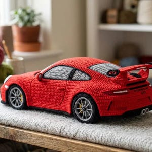 May include: A red crocheted sports car model featuring a rear spoiler, grey windows, and black trim. The rear of the vehicle displays the text Porsche GT3. The wheels are grey with yellow brake calipers, and the body is crafted from red yarn.