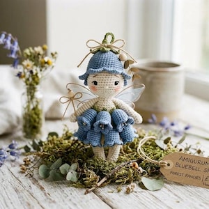 May include: A handmade amigurumi bluebell sprite doll with a blue hat and dress, and delicate wings. The doll is standing on a bed of moss, with a tag that reads "Amigurumi Bluebell Sprite". The background includes a vase of flowers.