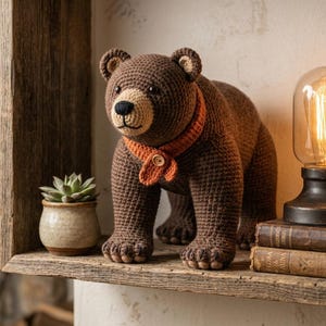 May include: A brown crocheted bear figurine featuring a tan muzzle, a black nose, and a textured orange scarf secured with a small wooden button. The bear is standing on four legs with visible paw details, crafted with a dense, uniform yarn stitch pattern.