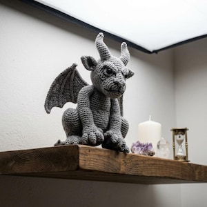 May include: A grey crocheted gargoyle figurine featuring pointed horns, textured wings, and a seated posture. The creature has a detailed face with a stern expression, crafted from yarn with visible stitches, positioned on a wooden shelf.