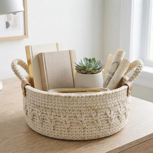 May include: A round, cream-colored crocheted basket with a textured cable pattern, featuring two sturdy handles attached with tan leather tabs. The basket contains two beige notebooks, a small succulent in a white pot, and a gold-toned pen.