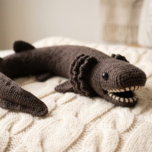 Puede incluir: Una criatura de ganchillo de color marrón oscuro que se asemeja a una anguila, con un cuerpo largo y curvado, aletas pequeñas y una boca ancha con dientes de color crema. El peluche tiene una superficie de hilo texturizado y un ojo de cuenta.