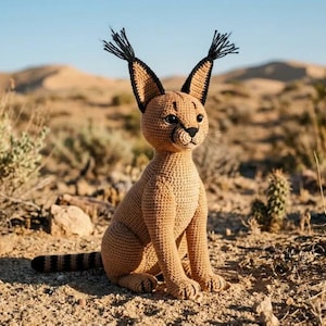 May include: A tan crocheted caracal plush toy featuring tall, pointed ears with black tufts at the tips, dark eyes, a small black nose, and a striped tail. The figure is crafted with a dense, textured yarn stitch pattern and sits upright.