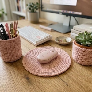 Puede incluir: Un conjunto de tres accesorios de escritorio tejidos a ganchillo en color rosa claro, que incluye un portalápices cilíndrico con varios bolígrafos y tijeras, una alfombrilla de ratón circular con un ratón rosa claro y una funda para maceta.