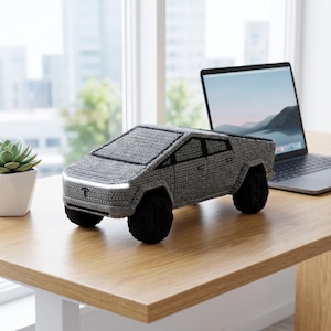 May include: A gray crocheted model of a pickup truck featuring black wheels, a white front light bar, and a small black logo on the front hood. The textile vehicle is crafted with visible yarn stitches and has a geometric, angular body design.