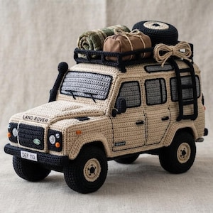 Puede incluir: Un juguete Land Rover Defender de ganchillo beige con detalles en negro, incluyendo una baca con una manta verde enrollada, una bolsa marrón y una rueda de repuesto. El vehículo cuenta con una escalera negra, ventanas grises y una matrícula DEF 110.