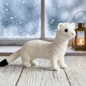May include: A white crocheted ferret toy with a long body, small rounded ears, and a black-tipped tail. The figure features dark round eyes and a small pink nose, crafted with visible yarn stitches in a standing pose on a white wooden surface.