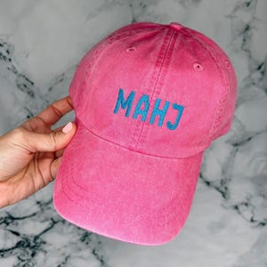 May include: A pink baseball cap with the word "MAHJ" embroidered in blue. The cap is made of a soft, textured fabric and has a curved brim. The hat is a vibrant pink color.