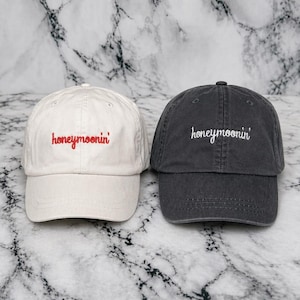 May include: Two baseball caps, one off-white and one charcoal gray, are displayed. The off-white cap has "honeymoonin'" embroidered in red, while the gray cap has the same text embroidered in white. Both caps are on a marble-patterned surface.