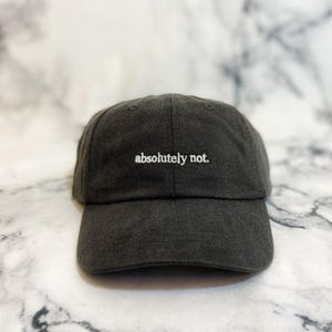 May include: A charcoal gray baseball cap featuring the text absolutely not. embroidered in white thread on the front center panel. The cap has a structured crown, a curved brim, and a fabric strap with a metal buckle closure at the back.