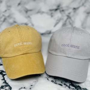 May include: Two baseball caps, one mustard yellow and one gray, are displayed. Each cap has the embroidered text "cool aunt" in a contrasting color. The caps are made of a soft fabric and have a curved brim.