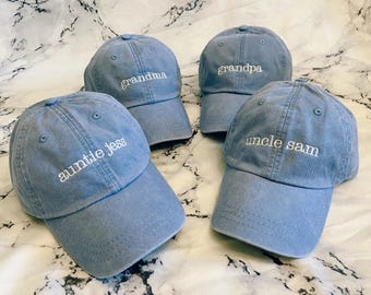 May include: Four blue baseball caps with white embroidered text. The caps read "grandma", "grandpa", "auntie jess", and "uncle sam". The caps are made of a soft material and have a curved brim.