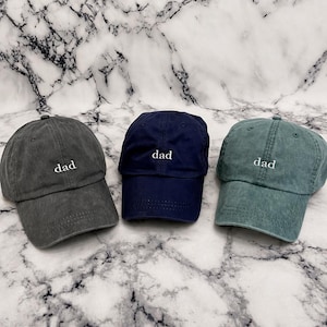 May include: Three baseball caps in charcoal gray, navy blue, and muted teal green. Each cap features a curved brim, a six-panel construction with eyelets, and the word dad embroidered in white lowercase letters on the front center panel.