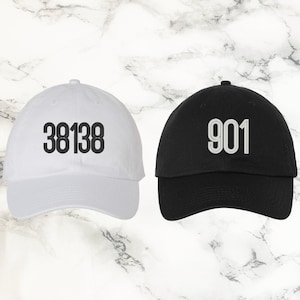 May include: Two baseball caps, one white and one black. The white cap has the number "38138" embroidered in black, while the black cap has "901" embroidered in white. Both caps are made of a soft fabric and have a curved brim.