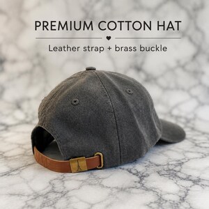 A gray cotton baseball cap with a leather strap and brass buckle, placed on a marble surface.