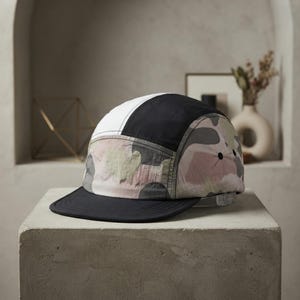 May include: A five-panel cap featuring a colour-blocked design with a solid white top panel, a solid black side panel, and a camouflage pattern in shades of muted pink, grey, and green. The cap has a flat, solid black brim and a clear plastic strap.