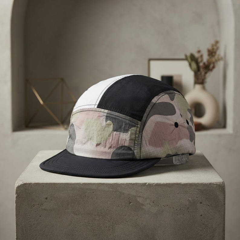 May include: A five-panel cap featuring a colour-blocked design with a solid white top panel, a solid black side panel, and a camouflage pattern in shades of muted pink, grey, and green. The cap has a flat, solid black brim and a clear plastic strap.