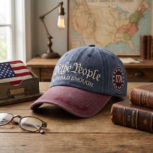 May include: A blue baseball cap with a maroon brim featuring the embroidered text We the People HAVE HAD ENOUGH in white on the front and a circular patch on the side with the number 1776 surrounded by stars, with additional text on the brim.