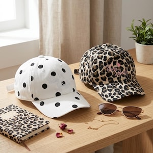 May include: Two baseball caps, one white with black polka dots and one with a brown and black leopard print. Both caps feature the embroidered text Ciao Bella. Also included are brown sunglasses, a leopard print notebook, and a gold necklace.