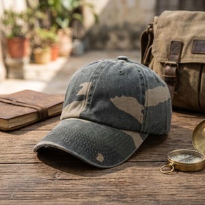 May include: A baseball cap featuring a washed-out camouflage pattern in shades of dark charcoal gray and muted beige. The cap has a curved brim, a six-panel construction with stitched eyelets, and a distressed finish on the fabric surface.
