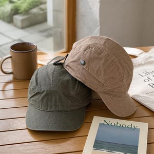 May include: Two five-panel caps, one in olive green and one in tan, featuring a structured crown, a short curved brim, and a small side button detail. The caps are made of a textured fabric and are positioned together on a wooden surface.