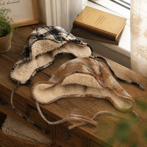 May include: Two trapper-style hats featuring a plaid pattern and a thick, cream-colored sherpa lining. One hat displays a black and white checkered design, while the other features a brown and blue plaid pattern. Both hats include long side ties.